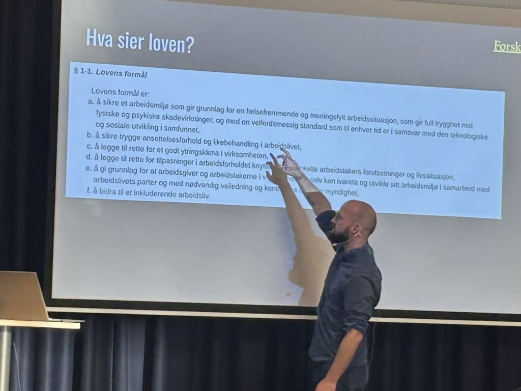 Klaus Lintho peker på en lysbildeskjerm med overskriften «Hva sier loven?» og teksten til arbeidsmiljøloven § 1-1.