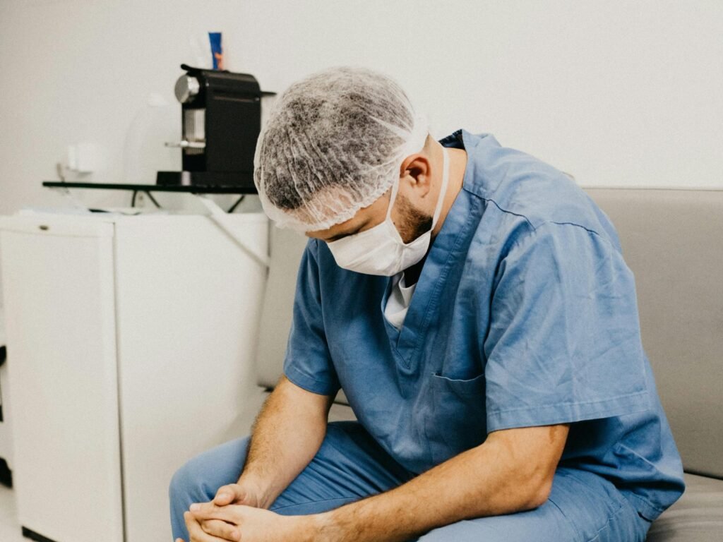 En helsearbeider i blå arbeidsklær og munnbind sitter foroverbøyd på en benk.